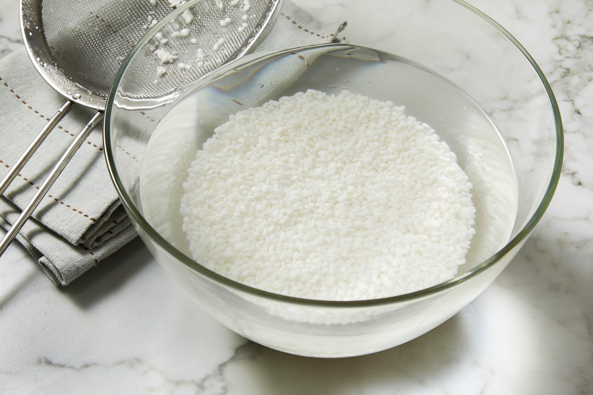 Overhead horizontal image of white short-grain rice soaking in water in a clear glass bowl.