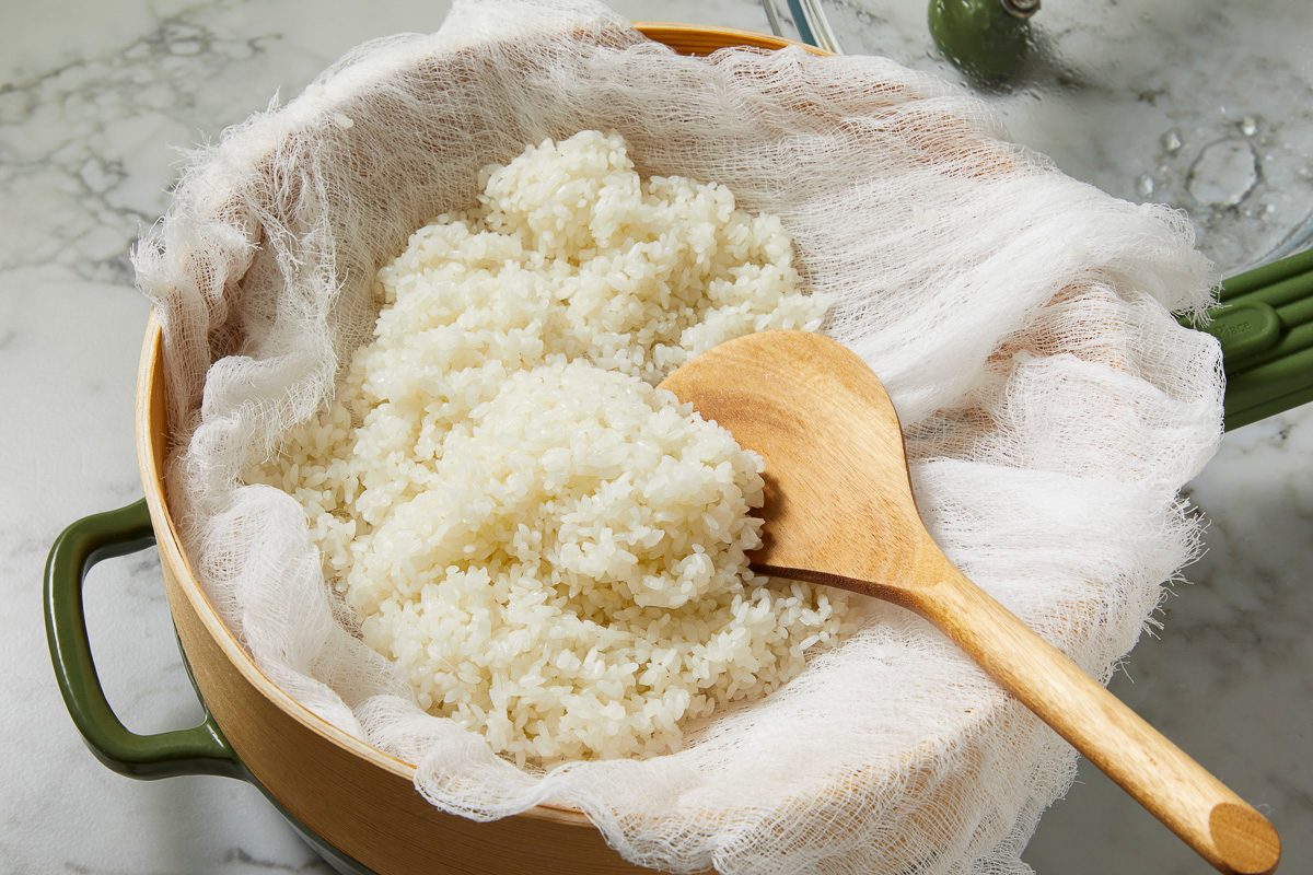 Fluffy, cooked white rice resting in a cheesecloth-lined bamboo steamer basket. A wooden spoon is placed in the rice.