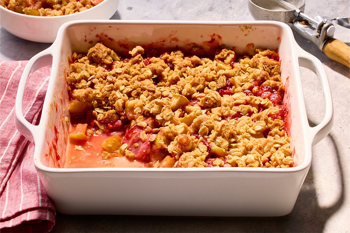 Overhead horizontal image of a freshly baked fruit crisp in a white rectangular baking dish. The crisp features a bubbly red fruit filling, likely containing apples and berries or rhubarb, topped with a golden-brown oat streusel. A red striped kitchen towel is to the left, and a metal ice cream scoop is visible to the right.