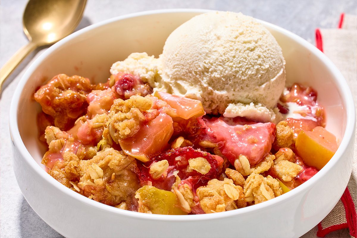 Eye level horizontal image of a serving of fruit crisp, likely apple and berry or rhubarb, in a white bowl. The warm fruit base is topped with a golden, oat-filled crumble and a large scoop of vanilla ice cream, with a gold spoon and linen napkin visible in the background.