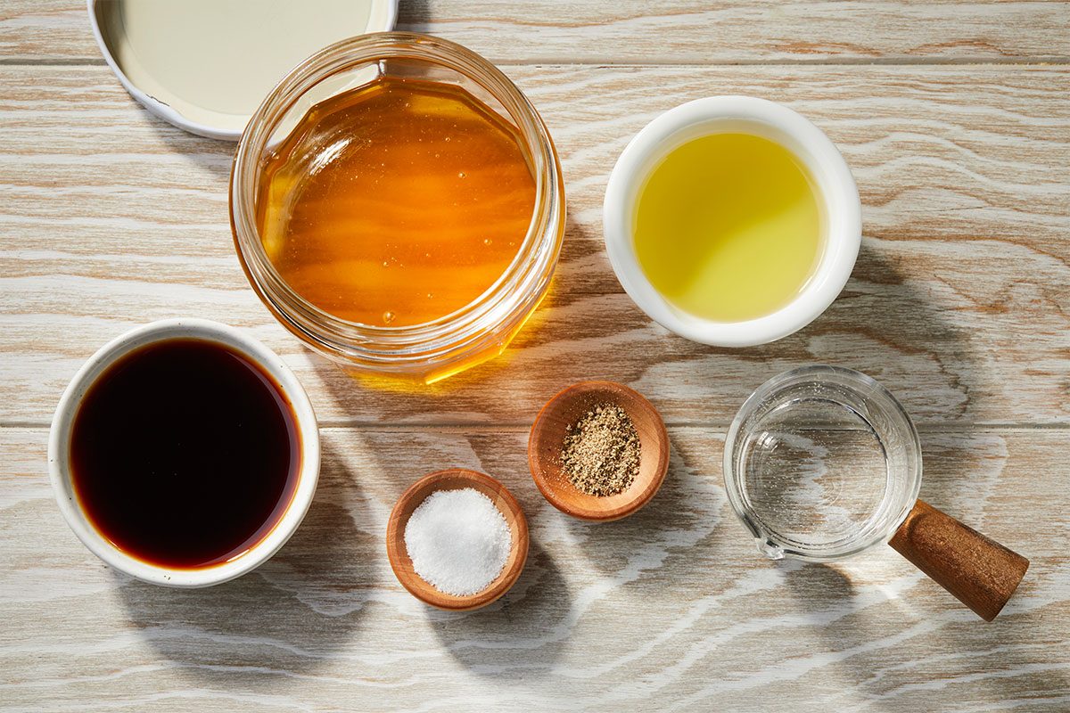 Overhead horizontal image of various dressing ingredients arranged on a light wooden surface. A jar of honey, bowls of olive oil and balsamic vinegar, and small dishes of salt and pepper are visible, along with a measuring cup of hot water. | [Original] RMS 274023 Strawberry Balsamic Pasta Salad; Trend Photography; How to; Step 1; Place honey, hot water, oil, vinegar, salt and pepper in a jar with a tight-fitting lid; shake well