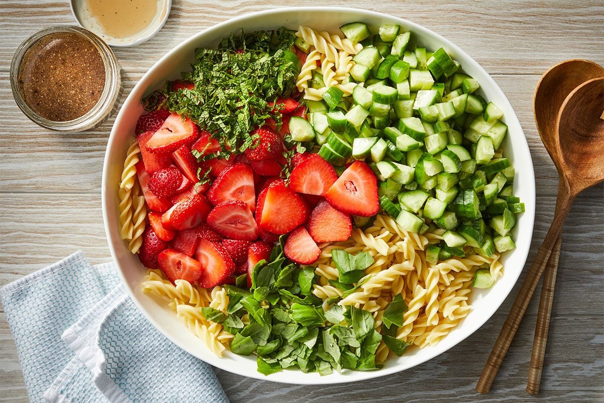 Overhead horizontal image of a large white bowl filled with ingredients for a salad. Fusilli pasta, chopped fresh strawberries, diced cucumbers, and fresh mint and basil leaves are neatly arranged in sections. A jar of dressing and wooden serving utensils are positioned beside the bowl on a light wooden surface. | [Original] RMS 274023 Strawberry Balsamic Pasta Salad; Trend Photography; How to; Step 2; n a large bowl, combine pasta, strawberries, cucumbers, mint and basil. Drizzle with dressing; toss to coat. Sprinkle with cheese