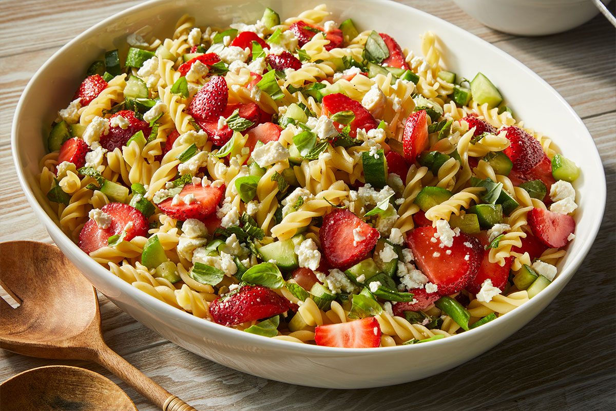 Eye Level horizontal image of a large white serving bowl filled with Strawberry Feta Fusilli Salad. The vibrant salad features fusilli pasta, chopped fresh strawberries, crumbled feta cheese, chopped cucumbers, and fresh green herbs like mint and basil. Wooden serving utensils are visible on the wooden table next to the bowl. | [Original] RMS 274023 Strawberry Balsamic Pasta Salad; Trend Photography; Horizontal AP; Pull