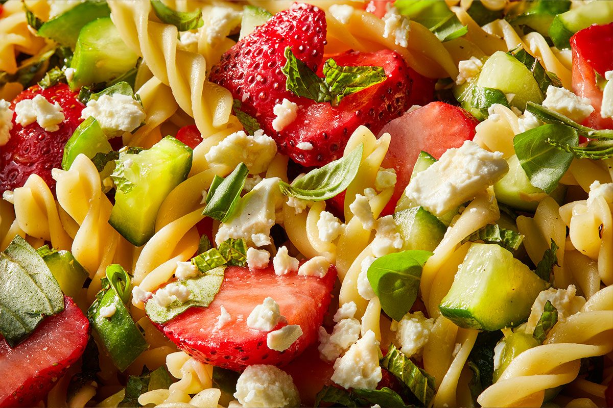 Overhead horizontal image of a vibrant Strawberry Feta Fusilli Salad. The close-up shot showcases tender fusilli pasta intertwined with bright red chopped strawberries, fresh green cucumber chunks, and crumbled white feta cheese. Specks of chopped fresh mint and basil leaves are scattered throughout the colorful mix. | [Original] RMS 274023 Strawberry Balsamic Pasta Salad; Trend Photography; Horizontal; Pull