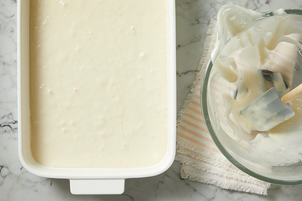 Overhead horizontal image of a white rectangular baking dish filled with a light-colored batter. To the right, a clear glass mixing bowl contains residual batter, with a gray silicone spatula resting inside. The items are placed on a white marble surface, next to a striped kitchen cloth.