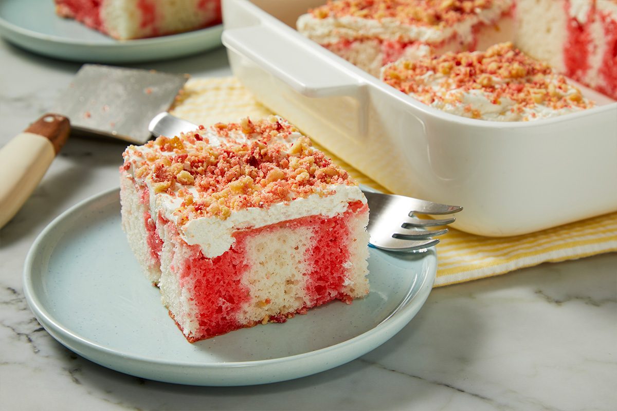 Eye Level horizontal image of a single slice of pink and white layered cake on a light blue plate with a fork. The cake is topped with fluffy white frosting and a golden and red crumble. In the background, a white baking dish holds more slices of the dessert.