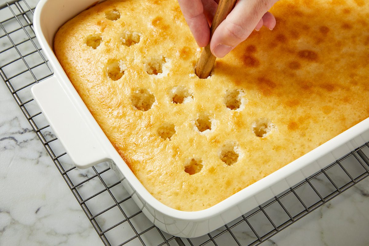 Eye level horizontal image of a hand using a wooden handle to poke holes into a baked cake. The golden-brown cake is in a white rectangular baking dish, and several holes are already visible across its surface.