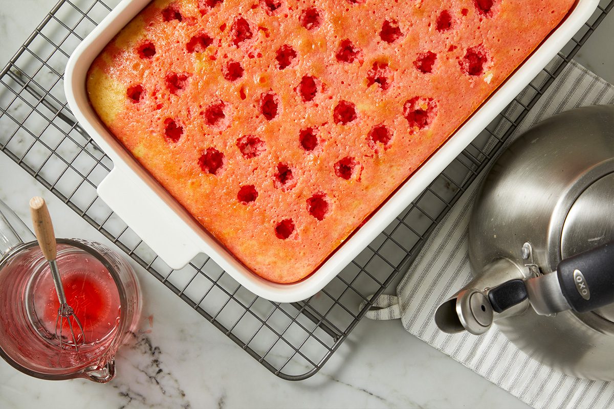 Overhead horizontal image of a baked poke cake in a white rectangular baking dish, resting on a wire cooling rack. The cake has numerous holes filled with a red liquid, and a glass measuring cup with more red liquid and a whisk, along with a silver kettle, are also visible on a marble surface.