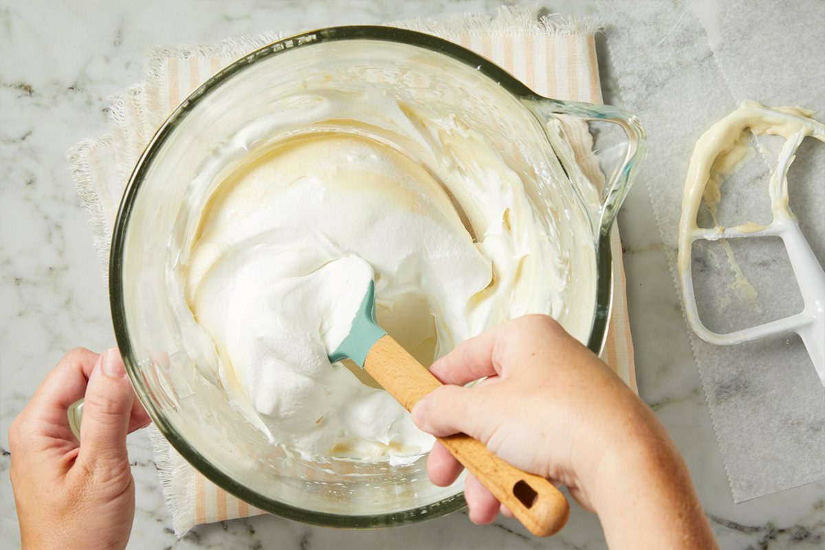 Overhead horizontal image of a hand using a spatula to gently fold a fluffy white mixture into a creamy yellow mixture within a clear glass bowl. A mixer paddle and a striped cloth are visible on the marble countertop next to the bowl.