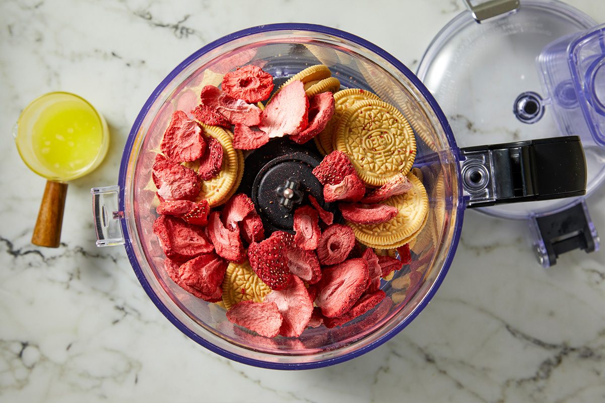 Overhead horizontal image of freeze-dried strawberries and golden sandwich cookies in the bowl of a food processor, with the lid open nearby. A small glass measuring cup containing melted butter is visible to the left of the food processor.