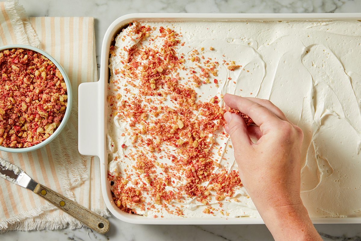 Overhead horizontal image of a hand sprinkling red and beige crumb topping over a white creamy layer in a white rectangular baking dish. A bowl of the crumb topping sits to the left, alongside a spreading knife on a striped napkin.
