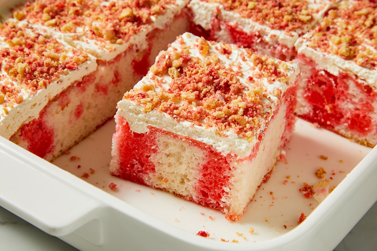 Eye Level horizontal image of sliced red and white marbled cake with white frosting and a crumble topping, served in a white rectangular baking dish. One slice is slightly separated from the rest, showcasing its vibrant layers and textured topping. The cake appears ready to be enjoyed, with a few crumbs scattered in the dish.