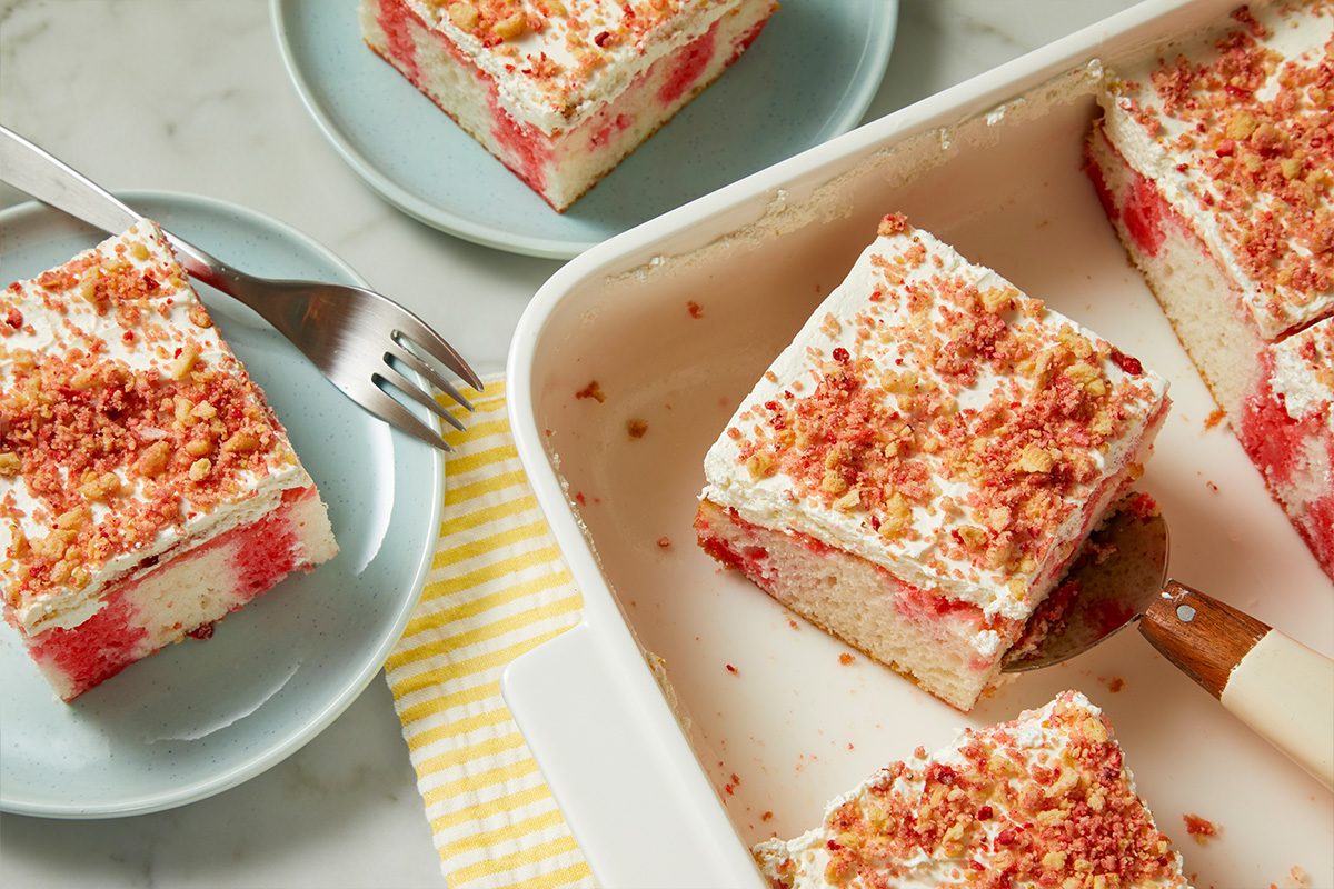 Overhead horizontal image of several slices of a white and pink swirled cake topped with whipped cream and strawberry crunch crumbs. One slice is being served from a white baking dish with a cake server, while other slices rest on light blue plates with a fork nearby.