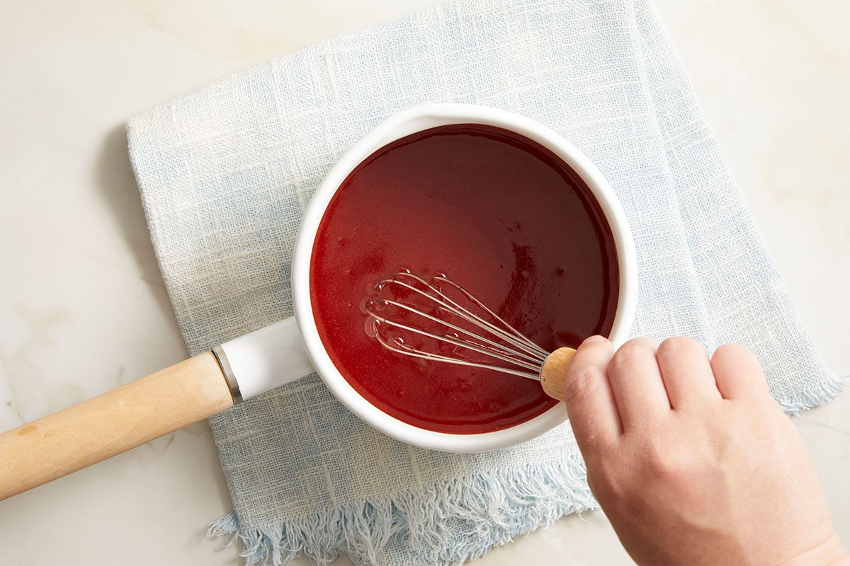 Overhead horizontal image of a hand whisking a dark red sauce in a white saucepan. The saucepan, featuring a wooden handle, rests on a light blue dishtowel. The scene is set on a light-colored marble surface, highlighting the process of combining ingredients.