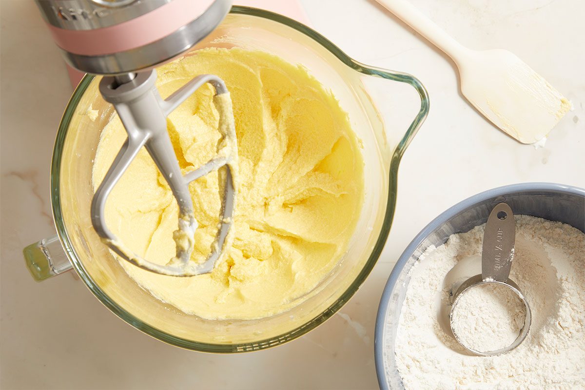 Overhead horizontal image of a pink stand mixer with a paddle attachment creaming a yellow batter in a glass mixing bowl. A bowl of white flour with a measuring cup and a spatula with some batter on it are visible on a white countertop.