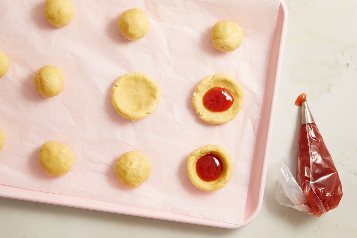 Overhead horizontal image of cookie dough balls on a pink baking sheet lined with parchment paper. Some of the dough balls are indented and filled with a red jam-like fruit filling. A piping bag containing more red filling is positioned on the right.