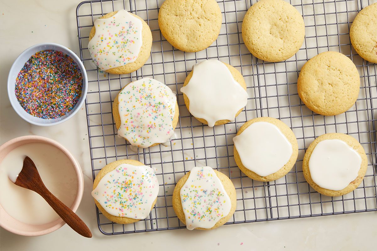 Overhead horizontal image of round cookies on a metal cooling rack. Some cookies are plain, while others are topped with white icing, some of which also have colorful sprinkles. A small bowl of white icing with a wooden spoon and another bowl filled with vibrant sprinkles are also visible.
