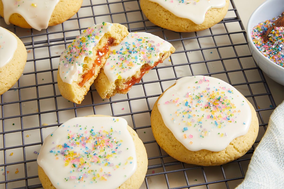 Overhead horizontal image of several round cookies with white icing and colorful sprinkles cooling on a wire rack. One cookie is broken in half, revealing a red jam filling inside. A white bowl filled with rainbow sprinkles is visible in the background.