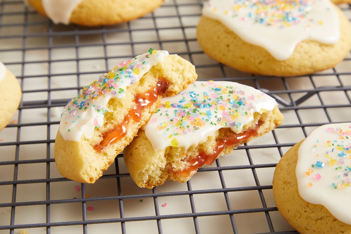 Eye level horizontal image of a jam-filled cookie broken in half, resting on a black cooling rack. The cookie halves reveal a vibrant red filling and are topped with white icing and colorful sprinkles, with additional cookies visible in the background.