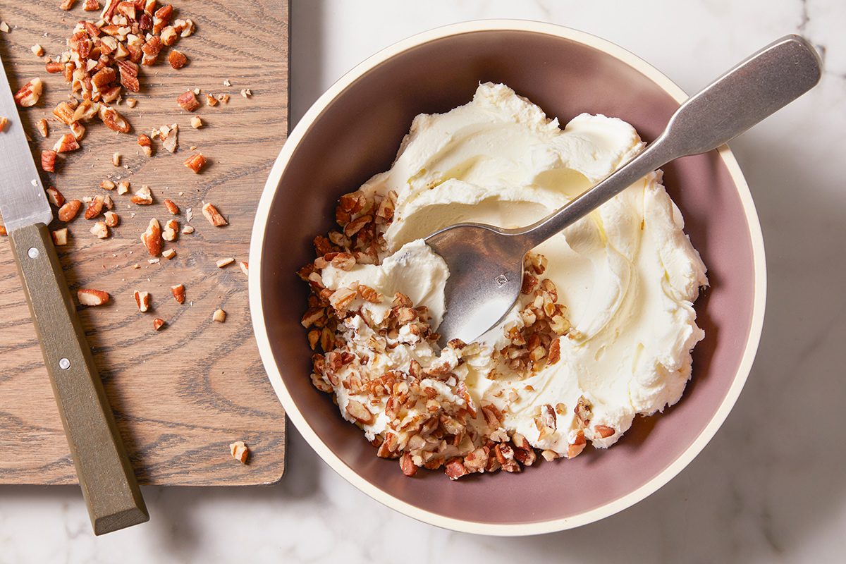 A bowl of yogurt topped with chopped nuts sits on a marble surface next to a wooden board with more chopped nuts and a knife. A spoon rests in the bowl.
