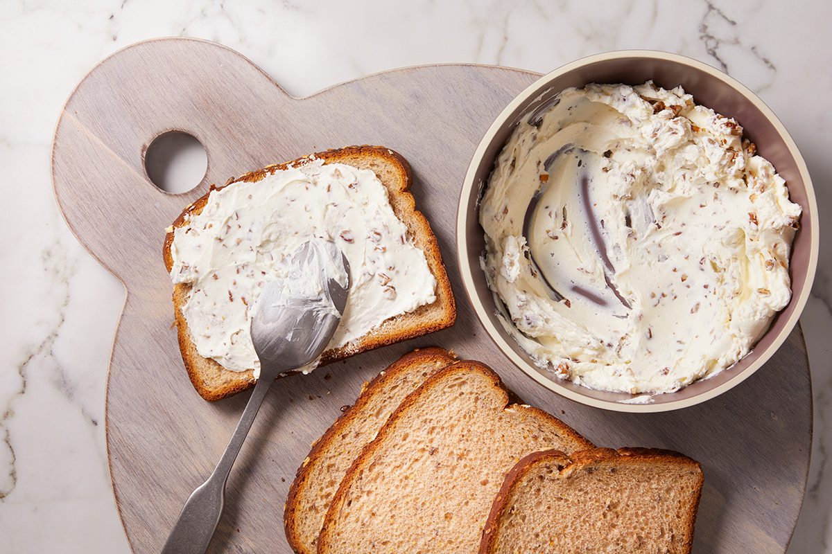 A slice of bread being spread with herbed cream cheese from a bowl, with additional plain slices nearby, all on a round wooden cutting board.