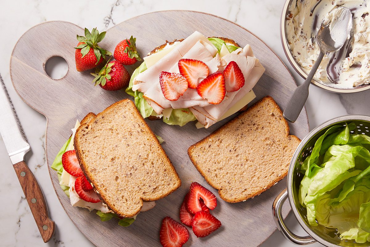 A sandwich with turkey, lettuce, and sliced strawberries on whole wheat bread sits on a round cutting board, surrounded by fresh strawberries, a knife, a bowl of lettuce, and a dish of cream cheese.
