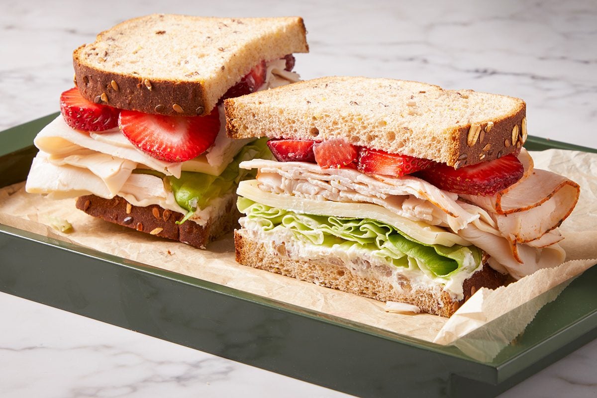 A sandwich made with whole grain bread, filled with layers of sliced turkey, leafy lettuce, and fresh strawberry slices, served on a green tray with parchment paper.