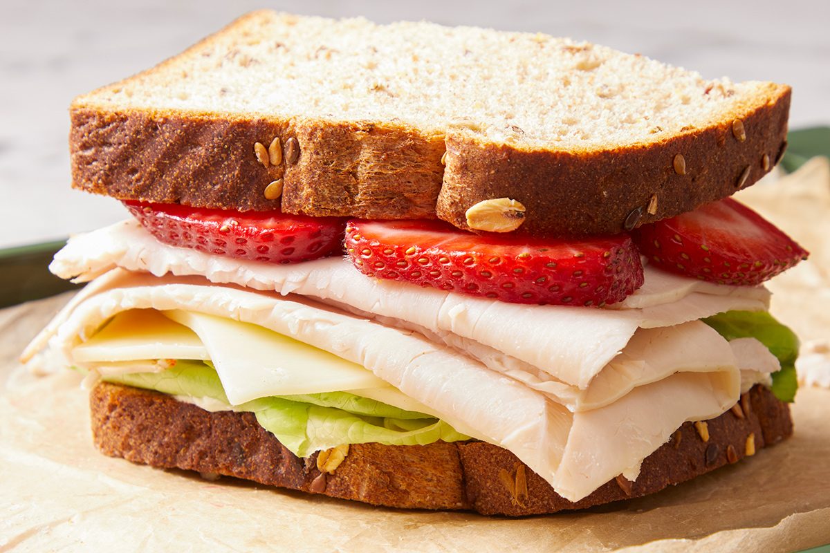 A sandwich made with whole grain bread, sliced strawberries, turkey, cheese, and lettuce, placed on parchment paper.
