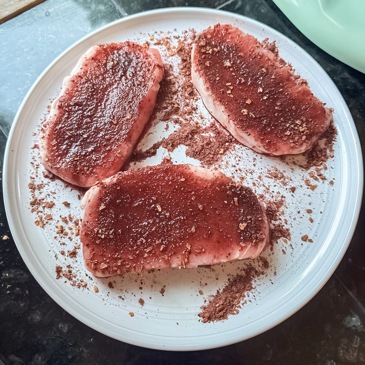 Three raw steaks coated with a reddish-brown spice rub are arranged on a white plate, with some excess spice scattered around them.