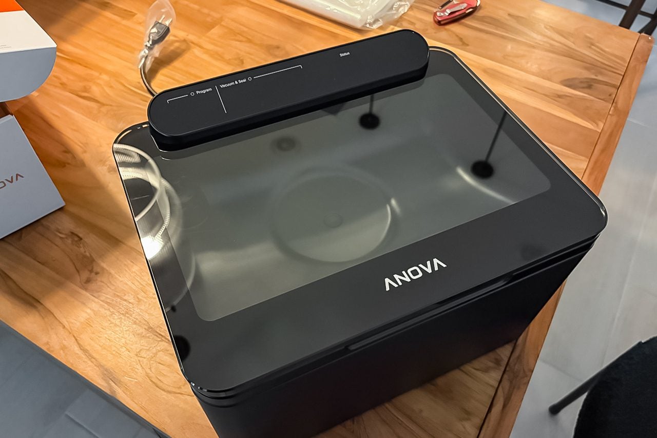 A black Anova sous vide cooker sits on a wooden table, viewed from above. The device has a transparent lid and a control panel on top.