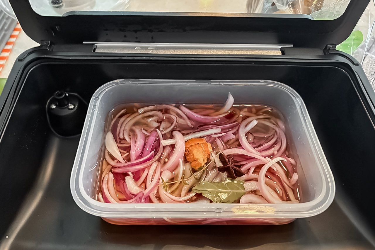 A plastic container holds sliced red onions, bay leaves, and a pepper soaking in liquid brine inside a black container with an open lid.