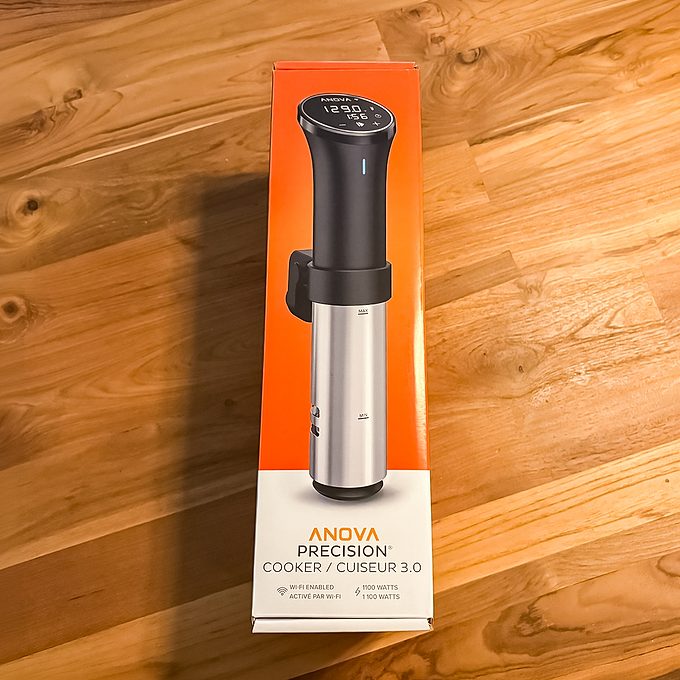 An Anova Precision Cooker 3.0 sous vide device in its packaging sits on a wooden surface. The box is orange and white with an image of the device and product details visible.