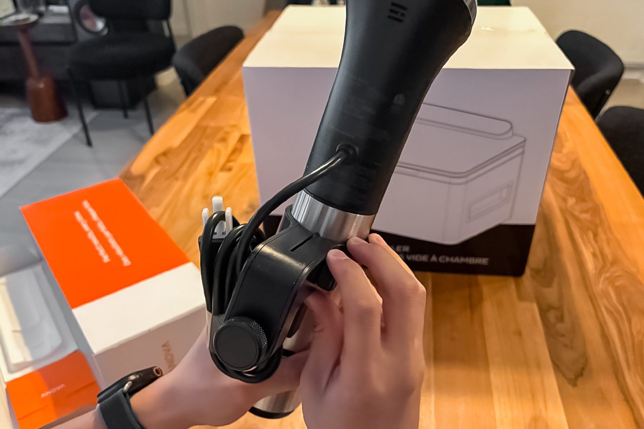 A person holds a black immersion circulator with a cord attached, above a wooden table with product boxes and kitchen chairs in the background.