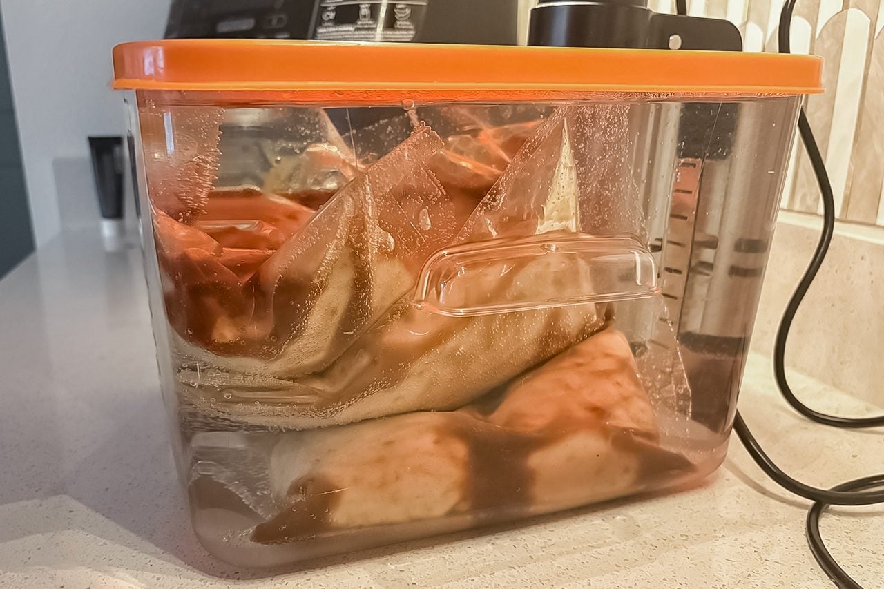A clear plastic container with an orange lid holds vacuum-sealed bags of food immersed in water, next to a sous vide machine on a kitchen counter.