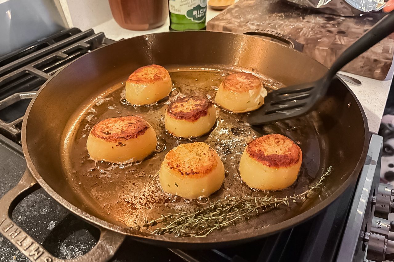 Six browned potato rounds are being cooked in a cast iron skillet with oil and herbs; a slotted spatula is turning one of the potatoes.