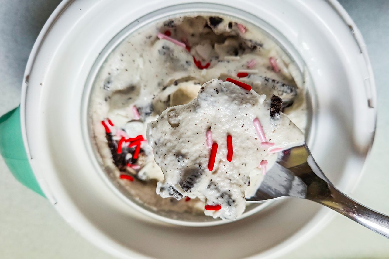 A close-up of a spoonful of cookies and cream ice cream with red and chocolate sprinkles, held above a bowl filled with more ice cream.