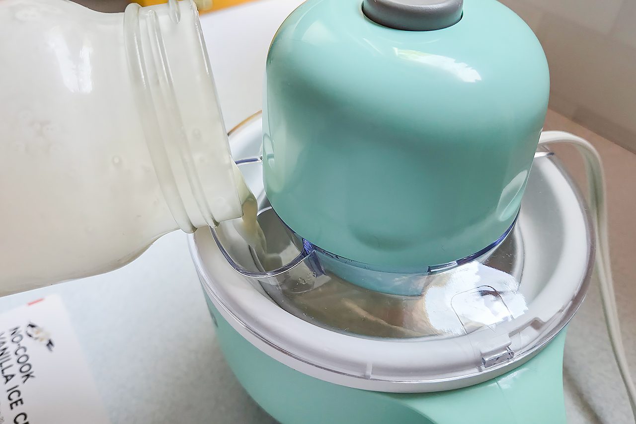 A jar pours liquid into a blue ice cream maker, preparing a homemade ice cream mixture. A recipe card is partially visible in the bottom left corner.