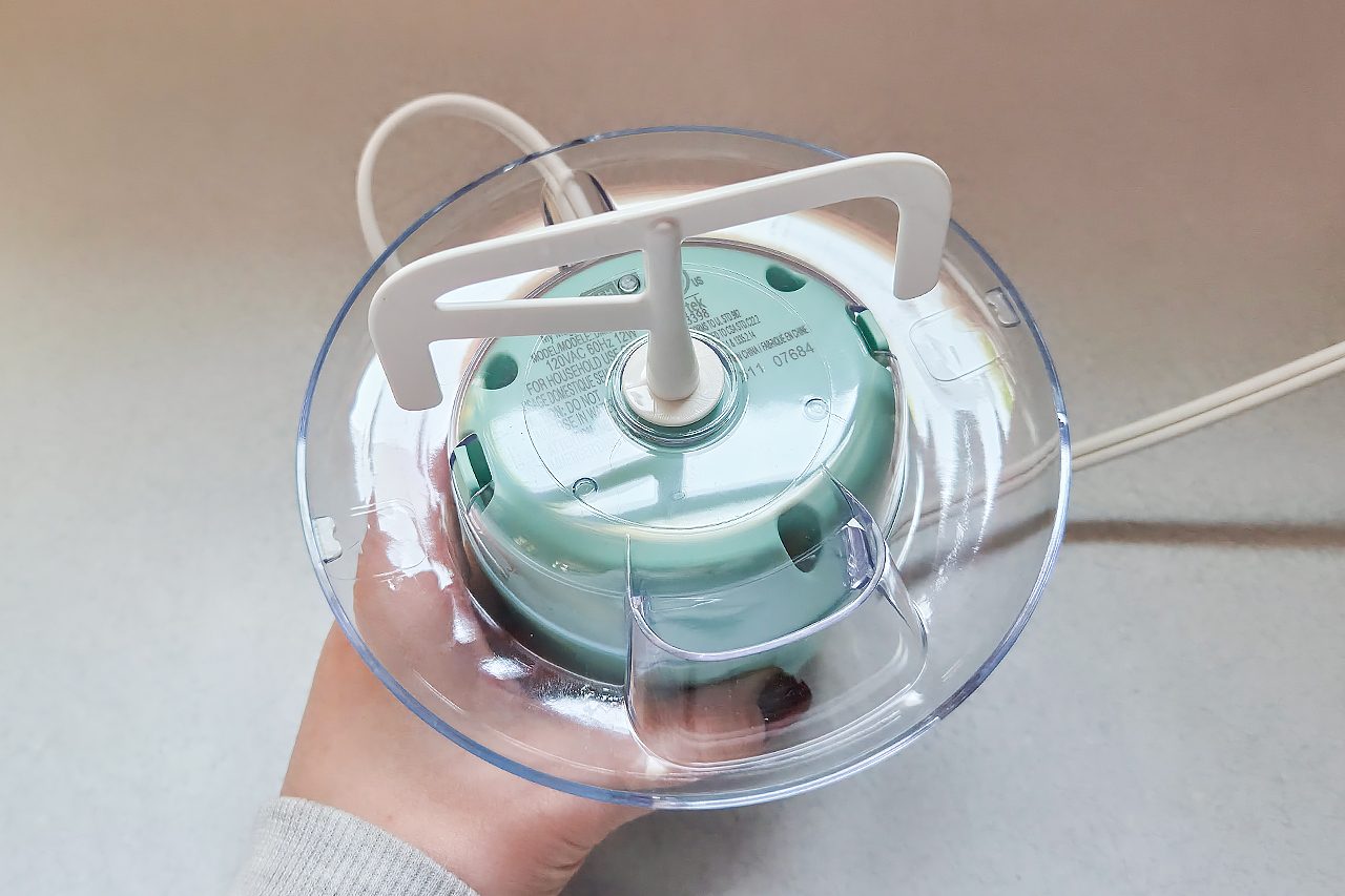 A hand holding the base of a small electric kitchen appliance with a clear plastic bowl and a white plastic spindle inside, along with a visible power cord.