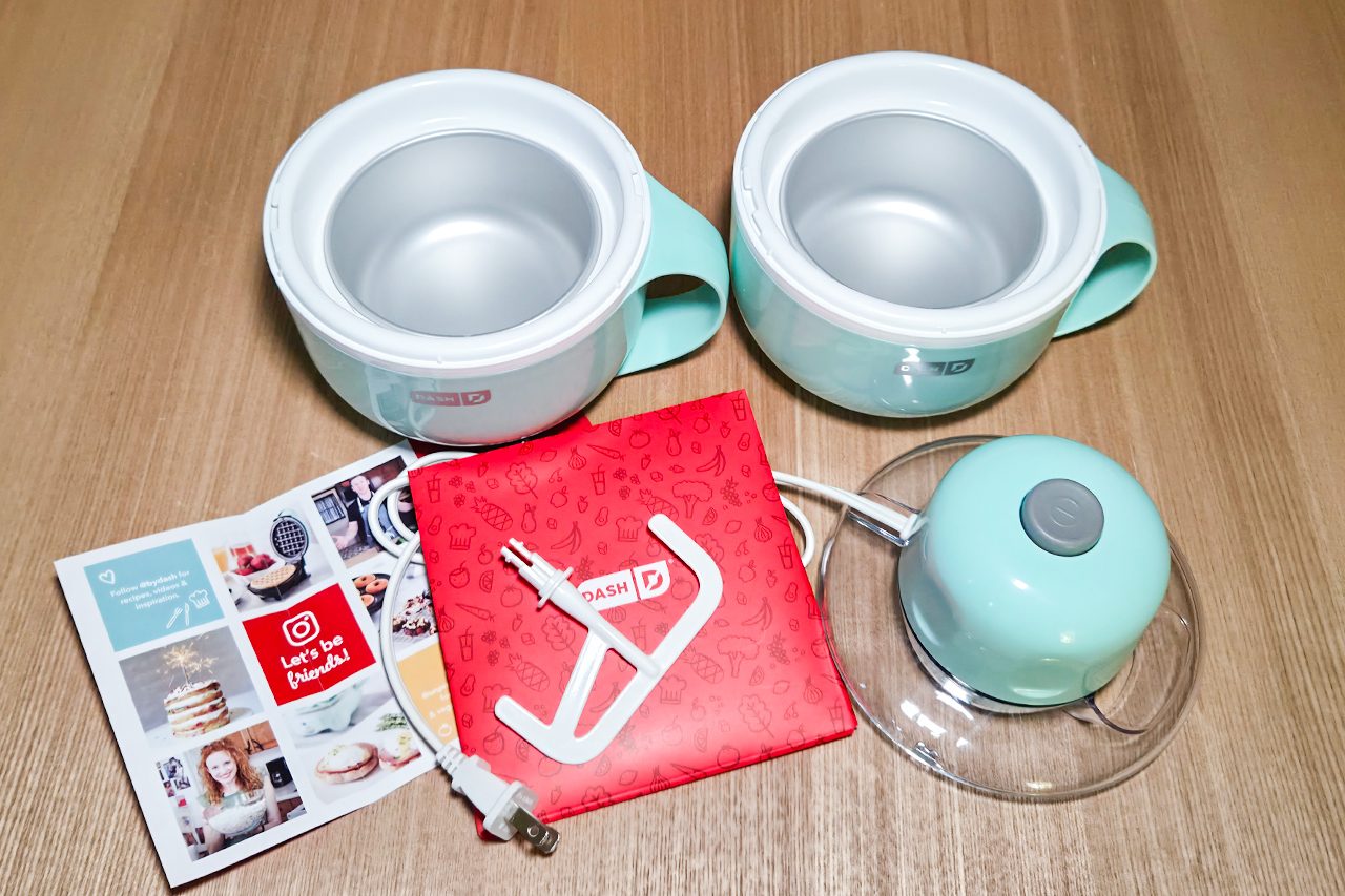 Two light blue mixing bowls, a power base with a grey button, a red instruction booklet, a white power cord, and a recipe booklet are arranged on a wooden surface.
