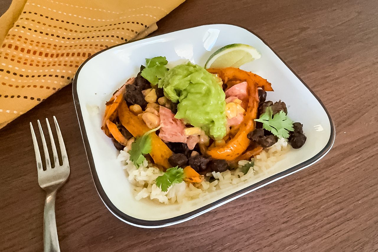 A square white bowl with rice, black beans, roasted sweet potato, shredded chicken, guacamole, cilantro, and a lime wedge, on a wooden table next to a fork and a yellow napkin.