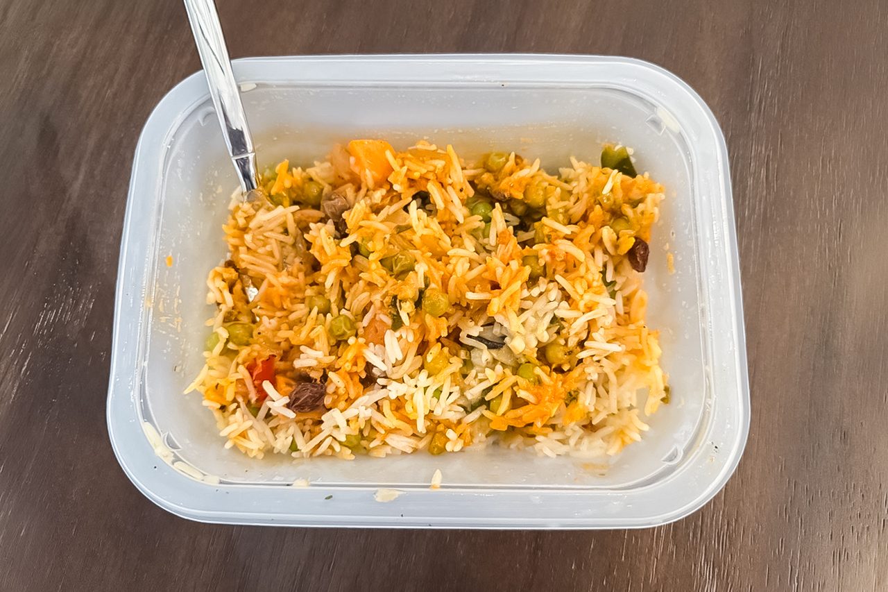 A plastic container filled with mixed rice, vegetables, and possibly raisins, with a metal spoon inside, placed on a brown wooden surface.