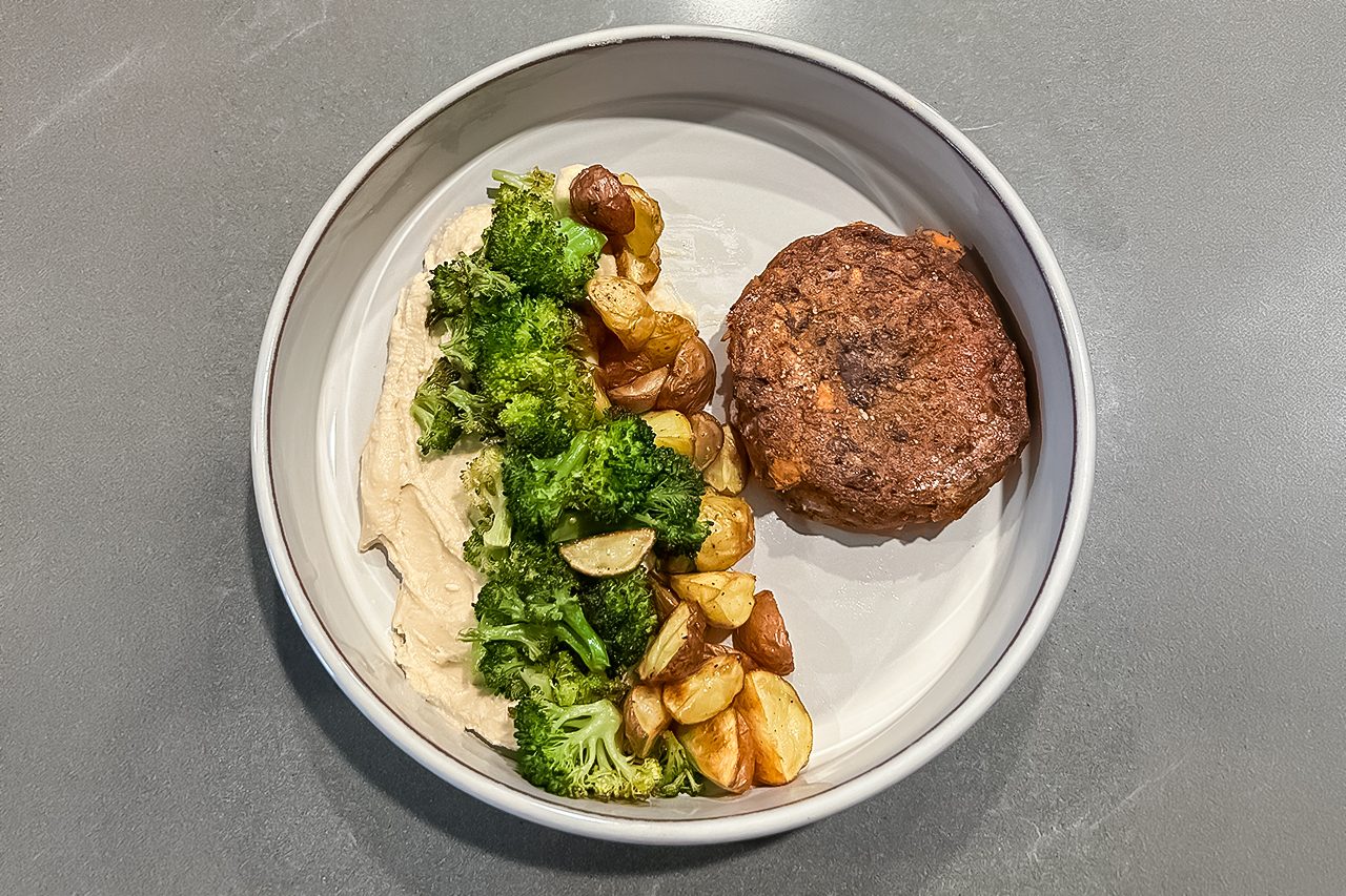 A plate with a round beef patty, roasted potatoes, broccoli, and a serving of mashed potatoes on a gray surface.