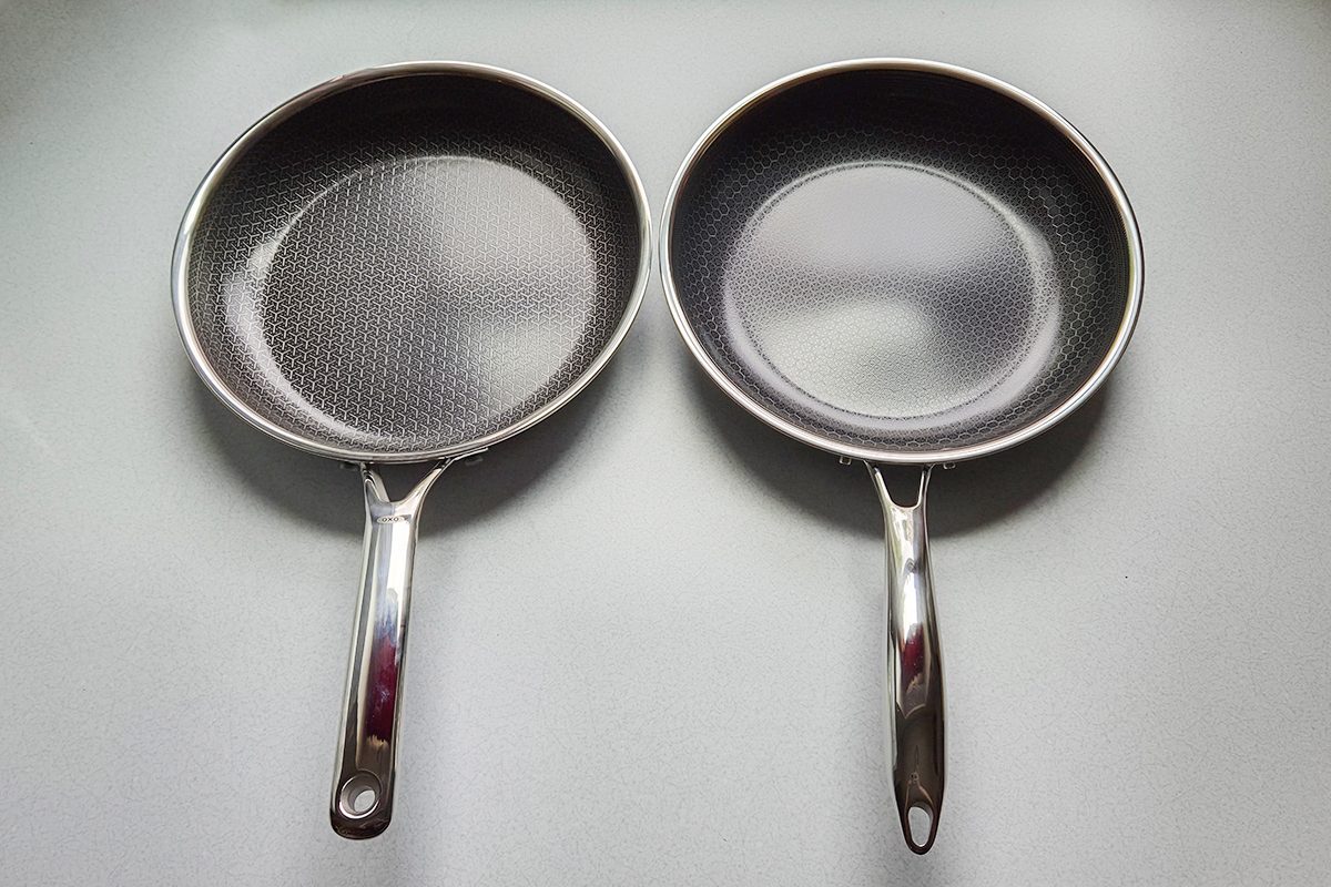 Two stainless steel frying pans with metal handles are placed side by side on a light-colored surface. The pans have a textured pattern on the inside.