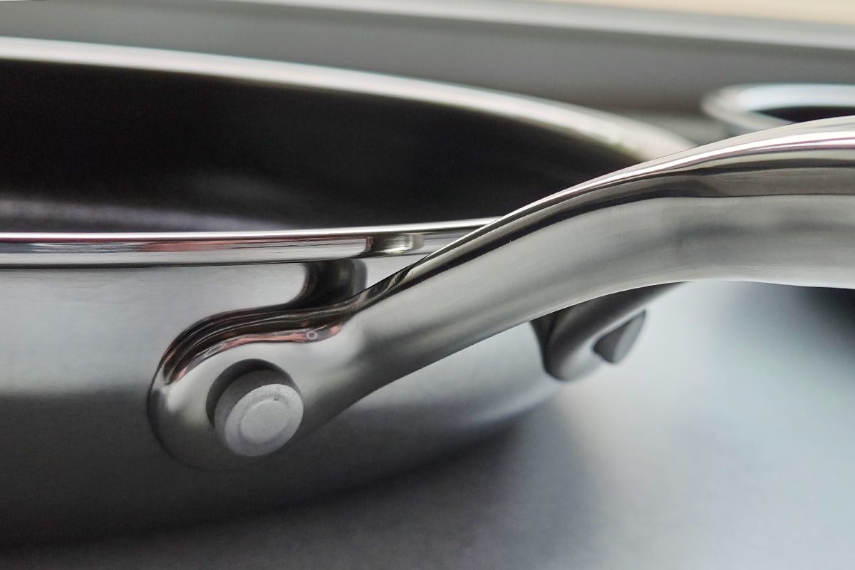 Close-up view of a stainless steel frying pan handle attached to the side of the pan, resting on a smooth gray surface.