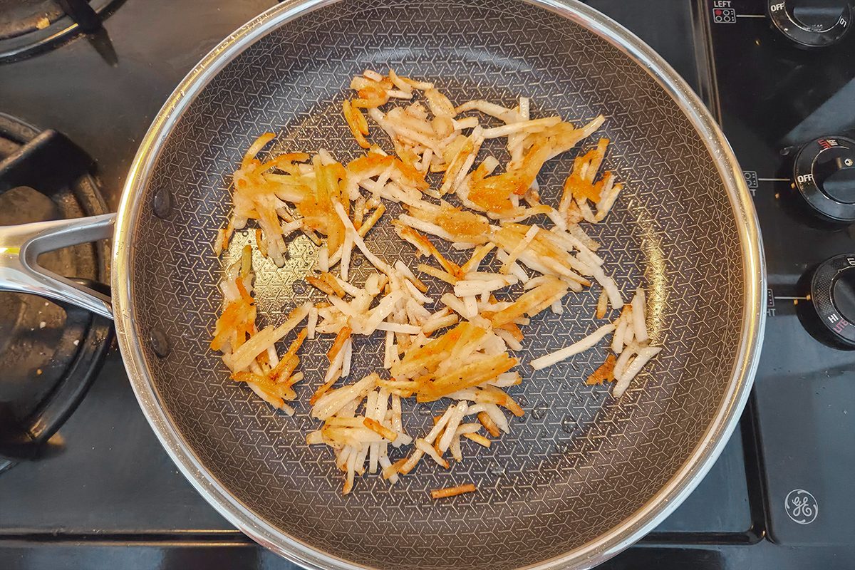 Shredded potatoes are being sautéed in a frying pan on a stovetop, showing some golden-brown edges.