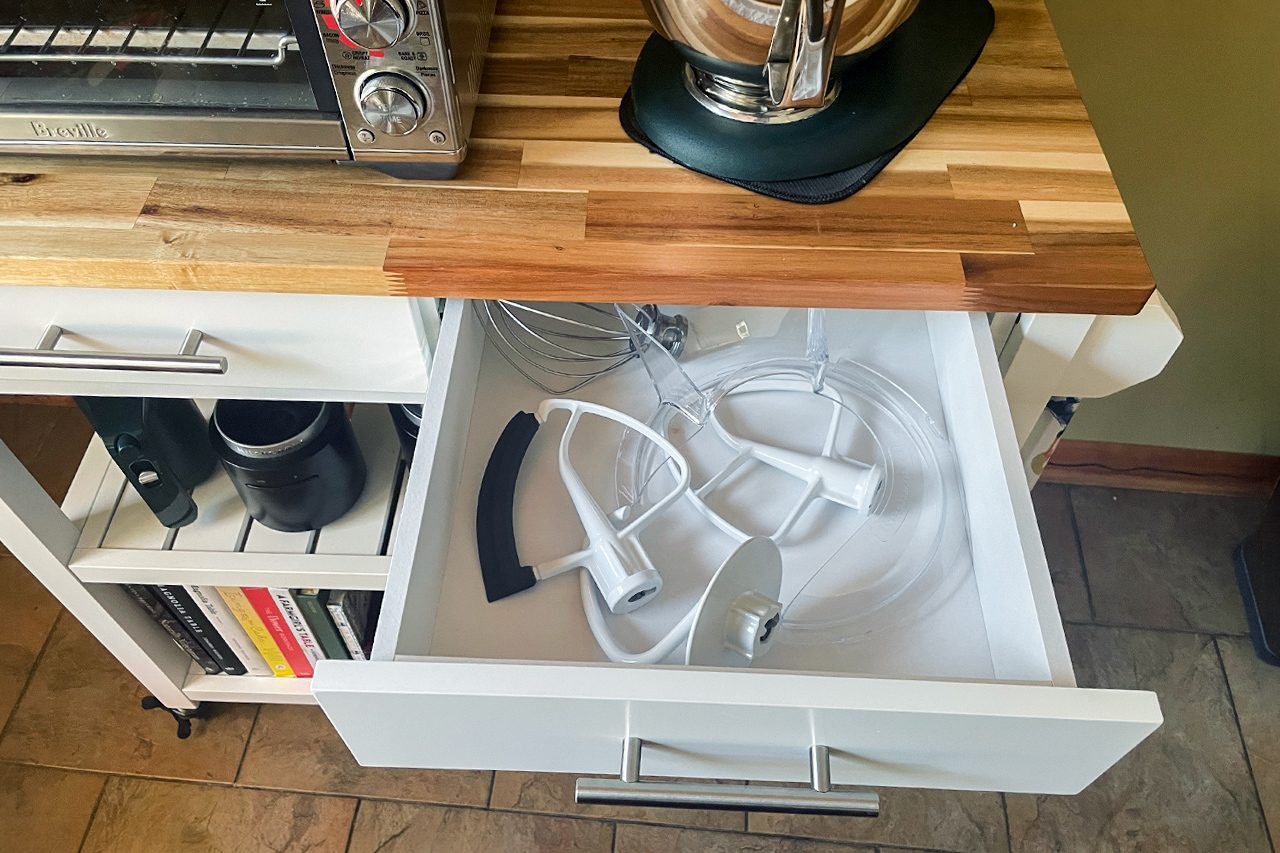 A kitchen drawer is open, showing several mixer attachments inside. Above the drawer is a butcher block countertop with a stand mixer and toaster on top. Shelves below hold mugs and books.
