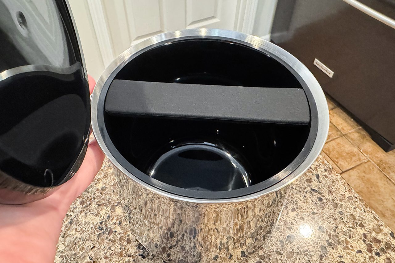 A hand holds open the lid of a stainless steel trash can on a granite countertop, showing a black foam insert inside the empty trash can.