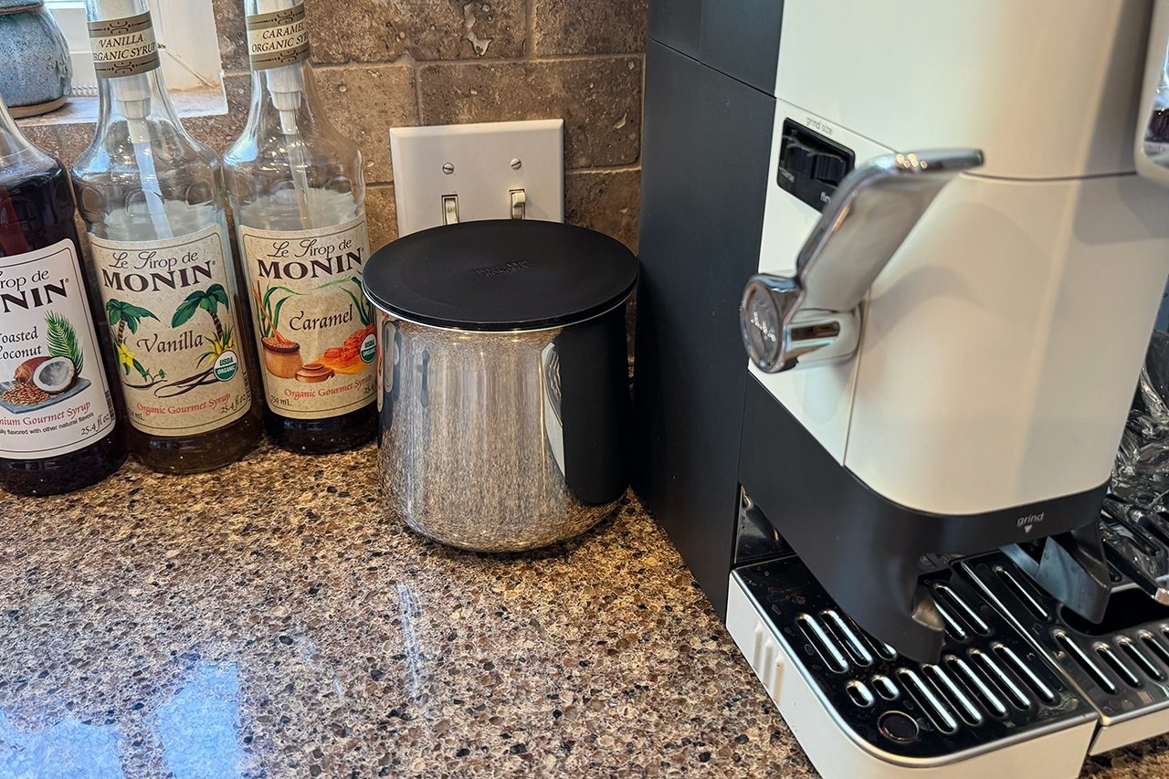 Bottles of Monin syrup, a metal canister with a black lid, and an espresso machine sit on a granite kitchen countertop near a tiled wall with outlets.