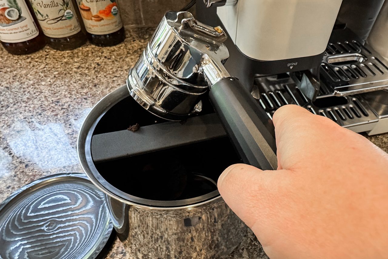 A hand taps a portafilter against a bar inside a knock box to remove used coffee grounds, with an espresso machine and bottles visible in the background.