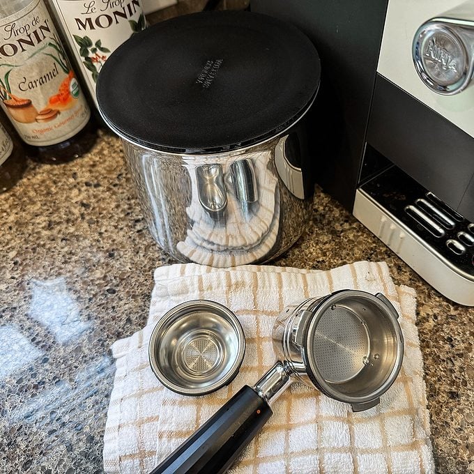 A portafilter, filter basket, and a metal knock box are on a checkered towel next to a coffee machine and Monin syrup bottles on a kitchen counter.